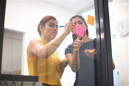 Two businesswomen writing out sticky notes on office glass wall Stock Photo - Premium Royalty-Free, Code: 649-08901446