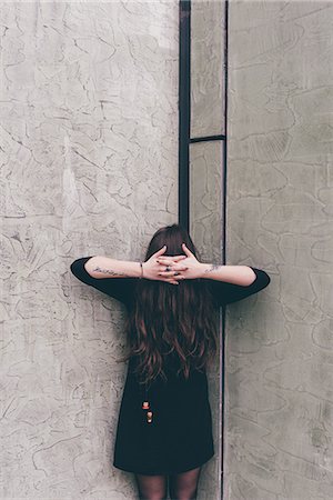 Portrait of young woman standing in corner, hair covering face, hands clasped in front of hair Stock Photo - Premium Royalty-Free, Code: 649-09025602