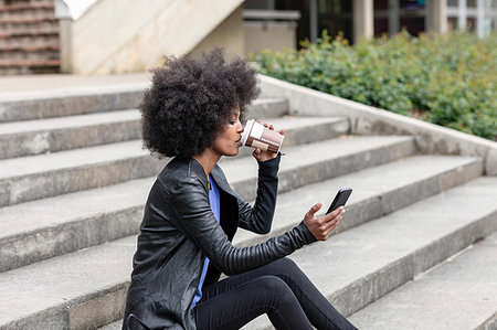 simsearch:614-09078761,k - Young woman with afro hair sitting on city stairway, drinking takeaway coffee and looking at smartphone Stock Photo - Premium Royalty-Free, Code: 649-09257705