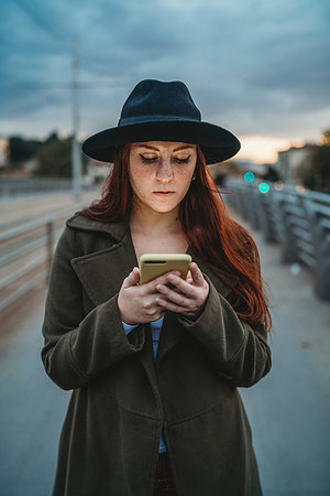 Young woman with long red hair on footbridge looking at smartphone at dusk Stock Photo - Premium Royalty-Free, Code: 649-09257914