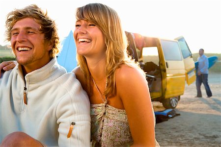 simsearch:649-01696405,k - Couple sitting on beach smiling with man carrying surfboard in background. Stock Photo - Premium Royalty-Free, Code: 649-01695949