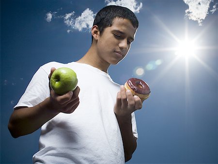 Low angle view of a teenage boy holding an apple and a donut Stock Photo - Premium Royalty-Free, Code: 640-01354860