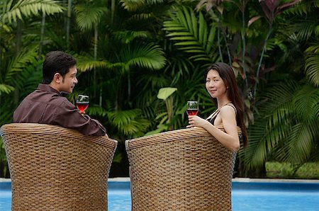 simsearch:841-02720357,k - Couple sitting by swimming pool in rattan chairs, woman turning to look at camera Stock Photo - Premium Royalty-Free, Code: 656-01765261