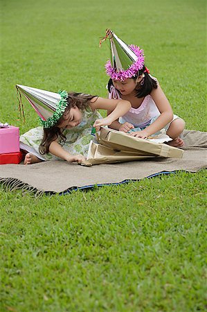 Two girls wearing party hats sitting on picnic blanket, opening a gift Stock Photo - Premium Royalty-Free, Code: 656-01765851