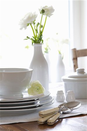 putting (placing object on or in something) - Stacked crockery and cutlery on a table with a vase of white ranunculus Stock Photo - Premium Royalty-Free, Code: 659-07027625