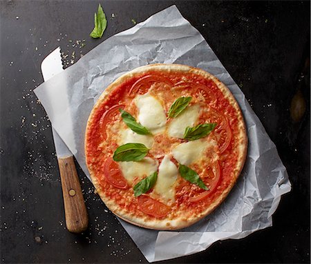 simsearch:659-08147216,k - A tomato, mozzarella and basil pizza on a piece of paper with a knife Stock Photo - Premium Royalty-Free, Code: 659-08147617
