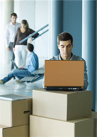 setup - Man using laptop on stack of cardboard boxes, colleagues talking in background Stock Photo - Premium Royalty-Free, Code: 695-05764171