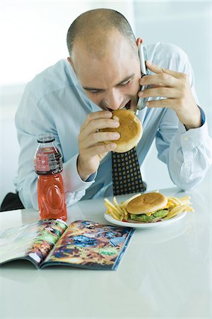 Man eating hamburgers, using cell phone, and looking at comic book Stock Photo - Premium Royalty-Free, Code: 695-05765624