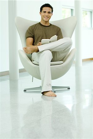 Young man sitting in chair holding book, smiling at camera, full length Stock Photo - Premium Royalty-Free, Code: 695-05767482