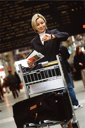 Woman standing with luggage cart in train station holding travel ticket looking at watch Stock Photo - Premium Royalty-Free, Code: 695-05769964
