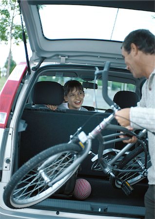 family loading car - Man putting bicycle into trunk of car, girl in car turning around smiling at man Stock Photo - Premium Royalty-Free, Code: 695-05777427