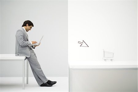 Businessman sitting on desk, shopping online, shopping cart and cursor in foreground Stock Photo - Premium Royalty-Free, Code: 695-05779422