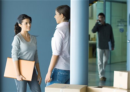 setup - Two young women standing face to face talking, man in background using cell phone Stock Photo - Premium Royalty-Free, Code: 695-03374282