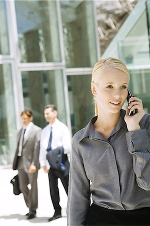 simsearch:695-03376228,k - Young businesswoman using cell phone outdoors, looking away, businessmen walking in background Stock Photo - Premium Royalty-Free, Code: 695-03376233