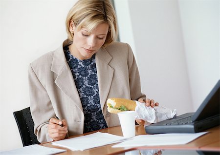 Businesswoman sitting at desk, holding sandwich and looking down at document Stock Photo - Premium Royalty-Free, Code: 695-03381725