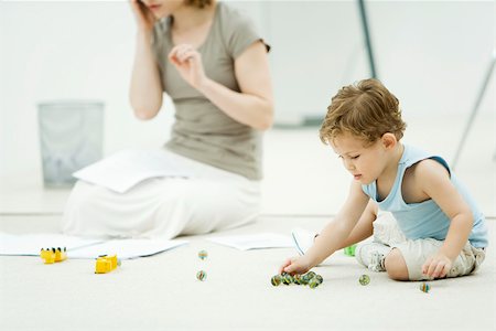 Little boy sitting on the ground, playing marbles, mother surrounded by paperwork in background Stock Photo - Premium Royalty-Free, Code: 695-03390034