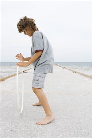 Boy standing on pier, playing with plastic hoop, side view, full length Stock Photo - Premium Royalty-Free, Code: 696-03402036