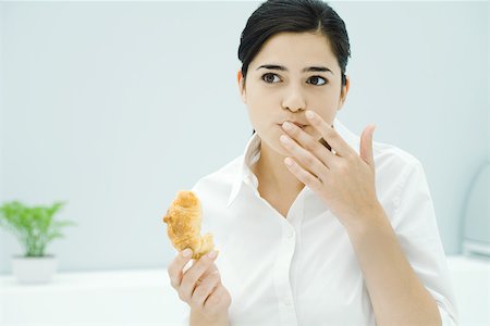Young woman holding croissant, other hand in front of mouth Stock Photo - Premium Royalty-Free, Code: 696-03402725
