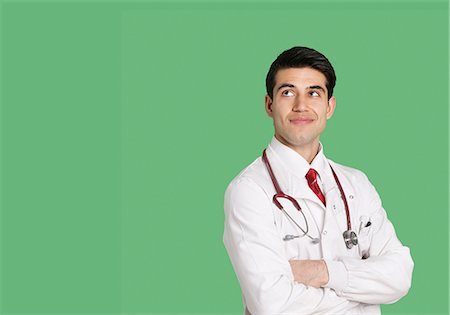 Male doctor in lab coat standing with arms crossed looking up over green background Stock Photo - Premium Royalty-Free, Code: 693-06324237