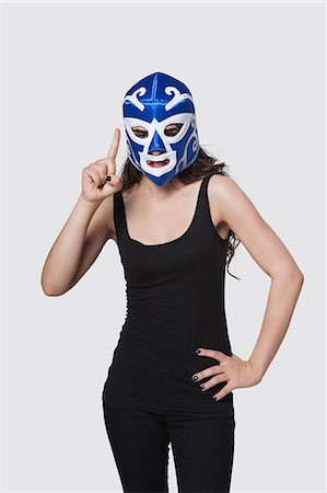 Young female wearing wrestling mask as she raises index finger against gray background Stock Photo - Premium Royalty-Free, Code: 693-06379882