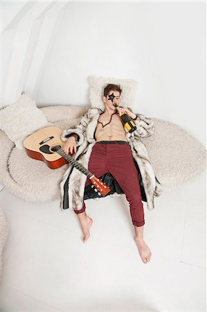 Young man with guitar drinking while lying on sofa Stock Photo - Premium Royalty-Free, Code: 693-06379933