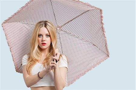 Portrait of young woman holding polka dotted umbrella against light blue background Stock Photo - Premium Royalty-Free, Code: 693-06379984