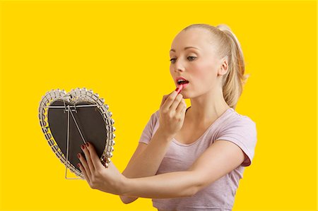 simsearch:693-06323962,k - Beautiful young woman looking at mirror while applying lipstick over yellow background Stock Photo - Premium Royalty-Free, Code: 693-06403286