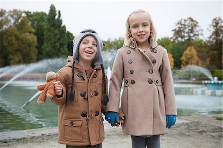 Portrait of happy brother and sister in trench coats holding hands at park Stock Photo - Premium Royalty-Free, Code: 693-06435947
