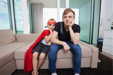 Portrait of smiling boy dressed in superhero costume sitting with sad father on sofa bed at home Stock Photo - Premium Royalty-Free, Code: 693-07455875