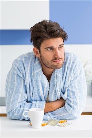 Young ill man with coffee mug and medicine leaning on kitchen counter Stock Photo - Premium Royalty-Free, Code: 693-07456338