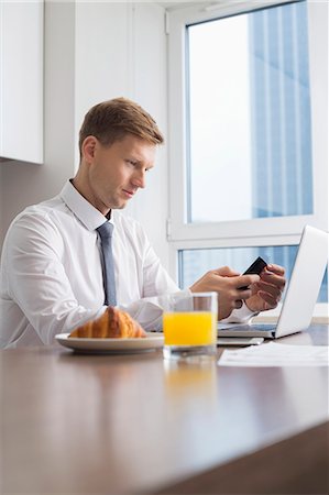 Mid adult businessman using cell phone with laptop on breakfast table Stock Photo - Premium Royalty-Free, Code: 693-07542229