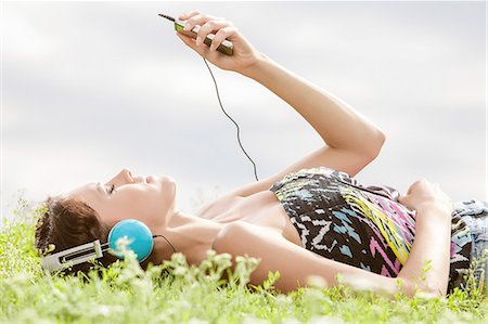 simsearch:693-07672615,k - Side view of young woman listening to music through MP3 player while lying on grass against sky Stock Photo - Premium Royalty-Free, Code: 693-07672806