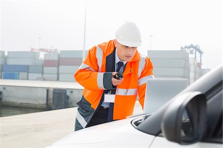 Middle-aged man talking on walkie-talkie while using laptop in shipping yard Stock Photo - Premium Royalty-Free, Code: 693-07913084