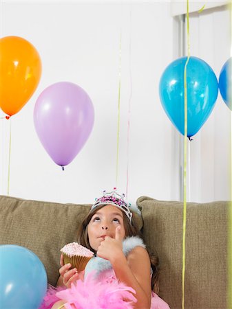 simsearch:693-03305634,k - Young girl (7-9) sitting on sofa eating cupcake, looking up at balloons Stock Photo - Premium Royalty-Free, Code: 693-03305629