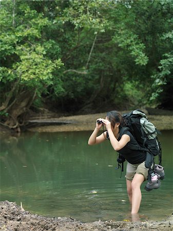 Young woman standing in water, carrying backpack, looking through binoculars Stock Photo - Premium Royalty-Free, Code: 693-03307518
