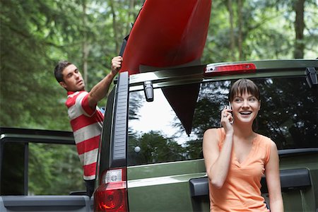 Woman using mobile phone by car, man tying kayak on roof, in forest Stock Photo - Premium Royalty-Free, Code: 693-03309077