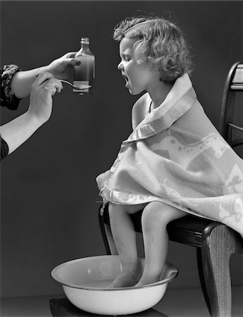 simsearch:846-05645854,k - 1940s SICK LITTLE GIRL SITTING IN CHAIR WRAPPED IN BLANKET FEET SOAKING IN HOT WATER TAKING MEDICINE ON SPOON FROM MOTHER Stock Photo - Rights-Managed, Code: 846-05646546