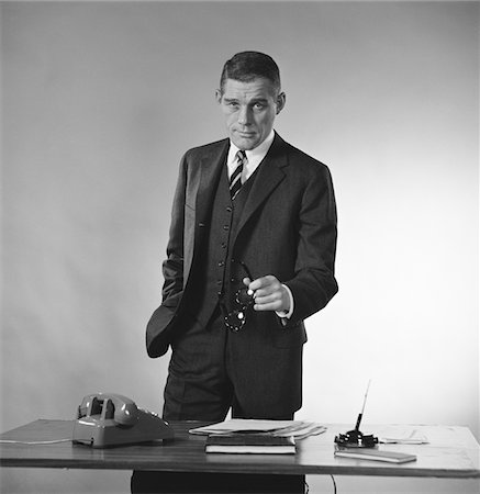 simsearch:846-05648290,k - 1960s SERIOUS BUSINESSMAN THREE PIECE SUIT STANDING BEHIND DESK GESTURING WITH GLASSES Stock Photo - Rights-Managed, Code: 846-05647745