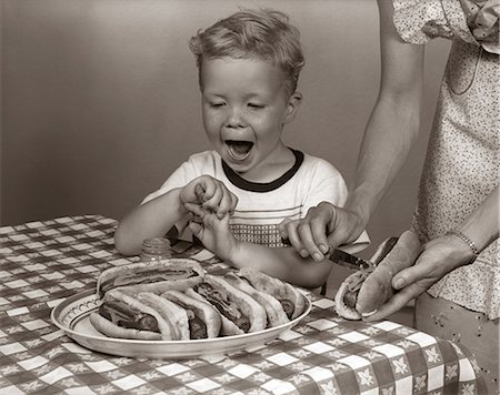 simsearch:846-05646226,k - 1950s EXCITED BOY AT TABLE WITH CHECKERED TABLECLOTH AND PLATTER OF HOT DOGS ON BUNS HIS MOTHER IS SPREADING MUSTARD ON ONE Stock Photo - Rights-Managed, Code: 846-06111797