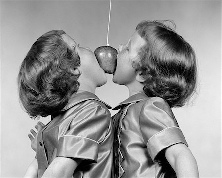 1950s IDENTICAL TWIN GIRLS TRYING TO EAT AN APPLE HANGING IN THE AIR FROM A STRING Stock Photo - Rights-Managed, Code: 846-06111880