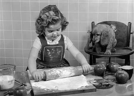 1950s LITTLE GIRL ROLLING OUT APPLE PIE CRUST ON KITCHEN TABLE WITH COCKER SPANIEL PUPPY WATCHING Stock Photo - Rights-Managed, Code: 846-06111985