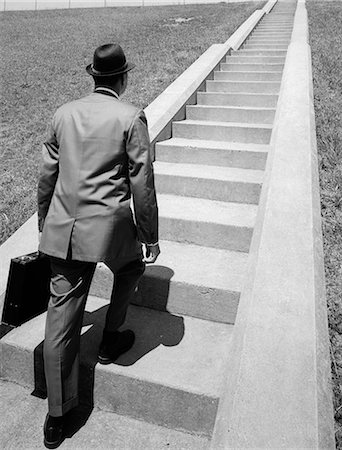 1960s BUSINESS MAN IN SUIT & FEDORA HOLDING BRIEFCASE STARTING UP LONG FLIGHT OF CEMENT STAIRS AHEAD OF HIM Stock Photo - Rights-Managed, Code: 846-06112444