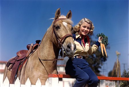 1940s 1950s SMILING YOUNG BLONDE COWGIRL SITTING ON FENCE POSING BY PALOMINO HORSE HOLDING BUNCH OF CARROTS LOOKING AT CAMERA Photographie de stock - Rights-Managed, Code: 846-07200117