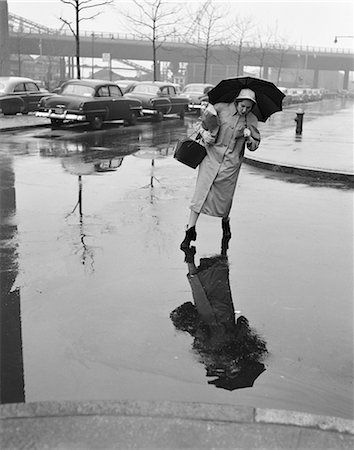 person stepping on a puddle - 1950s WOMAN IN RAIN COAT HAT BOOTS HOLDING UMBRELLA HANDBAG STEPPING TO AVOID PUDDLES WHILE CROSSING CITY STREET IN FOUL WEATHER Stock Photo - Rights-Managed, Code: 846-07200142