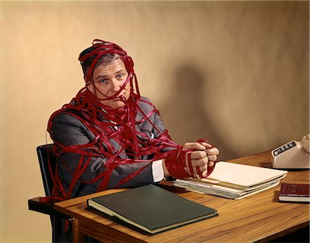 simsearch:846-08639518,k - 1960s BUSINESSMAN WRAPPED IN RED TAPE SITTING AT DESK WITH HANDS ON TOP OF STACK OF PAPER FOLDERS LOOKING AT CAMERA Stock Photo - Rights-Managed, Code: 846-07760741