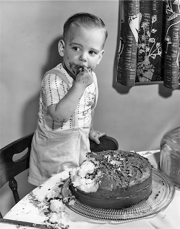 1950s LITTLE BOY TODDLER STANDING ON CHAIR BY CHOCOLATE CAKE WITH FINGERS IN MOUTH AND ICING ALL OVER HIS FACE Stock Photo - Rights-Managed, Code: 846-08721077