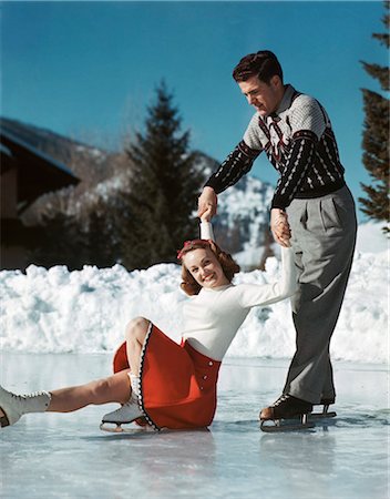 1940s 1950s COUPLE ICE SKATING SMILING WOMAN LOOKING AT CAMERA HAS SLIPPED AND FALLEN MAN HOLDING HER HANDS IS LIFTING HER UP Photographie de stock - Rights-Managed, Code: 846-09013087