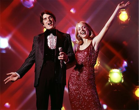 simsearch:846-05646372,k - 1970s WOMAN AND MAN WITH MICROPHONE SINGING AND GESTURING STROBE LIGHTS IN BACKGROUND TUXEDO GOWN ENTERTAINING Photographie de stock - Rights-Managed, Code: 846-09085345