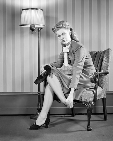 1940s FASHIONABLY DRESSED WOMAN BLOND VICTORY ROLLS HAIR STYLE LOOKING AT CAMERA SITTING ON LIVING ROOM CHAIR RUBBING SORE FOOT Foto de stock - Con derechos protegidos, Código: 846-09181501