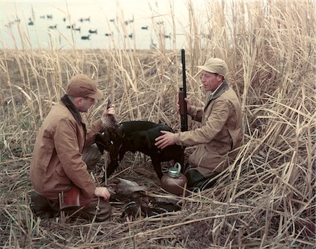 1950s TWO MEN BLACK DOG RETRIEVER IN TALL GRASS REEDS AT EDGE OF LAKE DUCK DECOYS AUTUMN WATER FOWL SPORT Stock Photo - Rights-Managed, Code: 846-02794184
